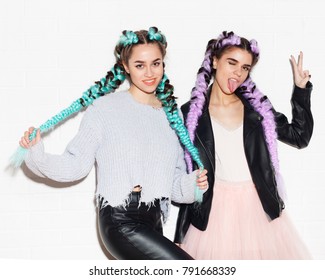 Two girls girlfriends with bright colored braids in fashion outfits having fun and show a sign of victory posing on white wall background