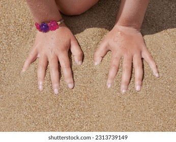 two girl's hands on wet white sand, pink and purple flower plastic bracelet on one hand - Powered by Shutterstock