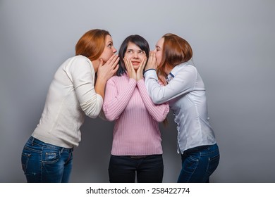 two girls in jeans European appearance whisper in the ear of the third girl in a sweater on a gray background, a secret girlfriend - Shutterstock ID 258422774