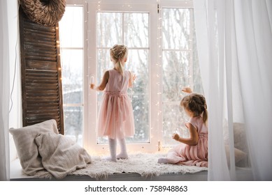 Two girls looking out the window at the winter landscape. Two little sisters in pink dresses waiting for christmas looking out of window. Waiting for the New Year.  - Powered by Shutterstock