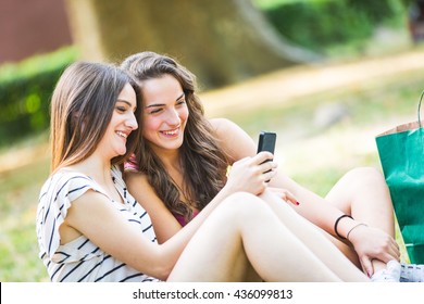 Two Girls Looking At Smart Phone At Park. They Are Happy And Smiling, Sitting On The Grass With Shopping Bags On Background. Friendship And Lifestyle Concepts.