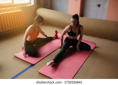 Two Girls On Sports Mats In A Fitness Center Stretch Their Inner Thigh Muscles, Pressing One Leg Under Themselves