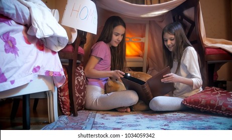 Two girls in pajamas sitting in selfmade tent in bedroom and reading big book