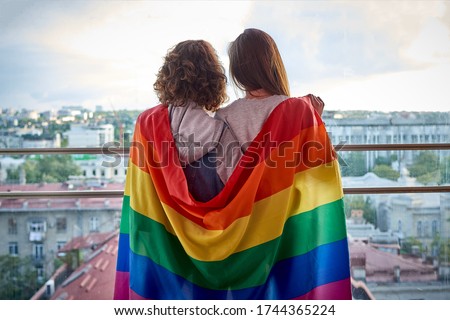 Two girls with rainbow flag looking out the window at the city. Lesbian couple celebrating pride month during Covid-19 quarantine, stay at home