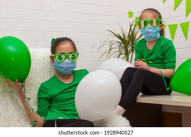 Two girls sisters in medical masks, dressed in green T-shirts holding balloons, having fun celebrating St. Patrick's Day.