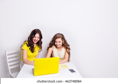 Two Girls Sit Behind A Laptop In Yellow Case. There Is A Smartphone On The Table Charging Via Yellow Cable. Both Girls Are Smiling And Looking At The Screen. One Wears Yellow T-shirt, Another - Dress.