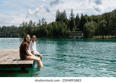 two girls sit on a wooden pier with their feet dangling in the clear blue water of the lake. They enjoy a tranquil atmosphere, surrounded by green trees and a wooded landscape as a backdrop. The sky i - Powered by Shutterstock