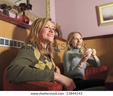Two girls are sitting in a cafe, laughing and drinkiing coffee.