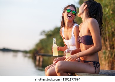 Two Girls Sitting And Having Fun On The Pier By The Lake On Sunset.