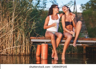 Two Girls Sitting And Having Fun On The Pier By The Lake On Sunset.