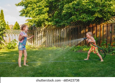 Two girls splashing each other with gardening house on backyard on summer day. Children playing with water outside at sunset. Candid moment, lifestyle activity. Life friendship of sisters siblings. 