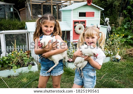 Two girls standing in front of hen house in a garden, holding white chickens, smiling at camera.