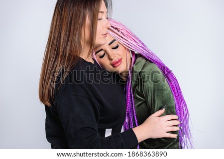 Two girls together indoors. A girl with pink hair braids lies on friend shoulder. White background.