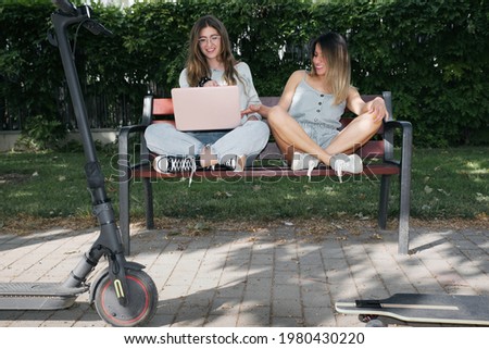 Two girls using the computer on a park seat, with an electric scooter and a skateboard.