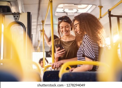 Two girls watching phone and smiling while standing on a bus.