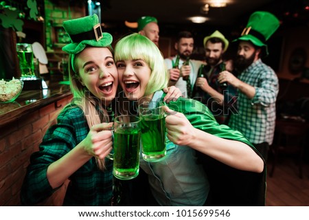 Two girls in a wig and a cap are photographed in a bar. They celebrate St. Patrick's Day. They are having fun.