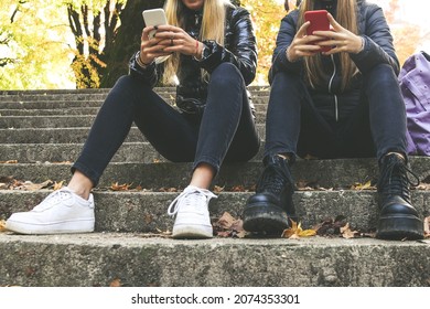 Two Girls With Wool Cap Sitting In The Park Using Smartphone, Beautiful Autumn Afternoon. Teen Using Mobile Phone. Sisters Chat With Friends And Classmates. Young Women Enjoying Free Time In Nature