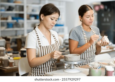 Two girls are working hard on creating ceramic products. Young employee of pottery workshop sculpts blank from clay, assistant applies drawing with brush and paints surface of ceramic vase. - Powered by Shutterstock