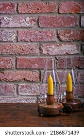 Two Glass Hurricane Candle Lamps Stand On Antique Wooden Table In Front Of Red Brick Hearth