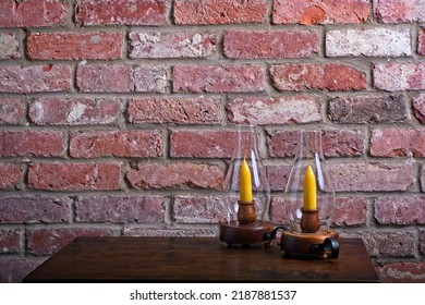 Two Glass Hurricane Candle Lamps Stand On Antique Wooden Table In Front Of Red Brick Hearth