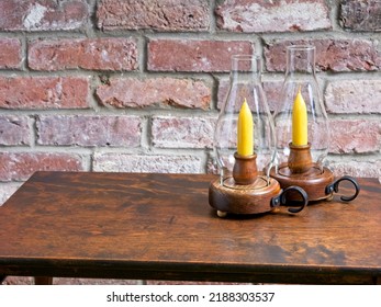 Two Glass Hurricane Candle Lamps Stand On Antique Wooden Table In Front Of Red Brick Hearth