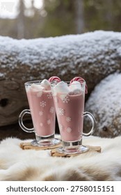 Two glass mugs filled with ruby chocolate hot chocolate topped with whipped cream, marshmallows and a candy cane. The snowflake mugs, wooden coasters and soft fur surface create a cozy, festive winter atmosphere with a distinctive pink ruby chocolate tone.