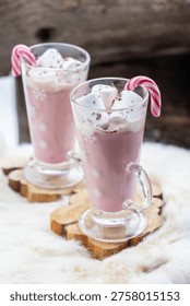 Two glass mugs filled with ruby chocolate hot chocolate topped with whipped cream, marshmallows and a candy cane. The snowflake mugs, wooden coasters and soft fur surface create a cozy, festive winter atmosphere with a distinctive pink ruby chocolate tone.