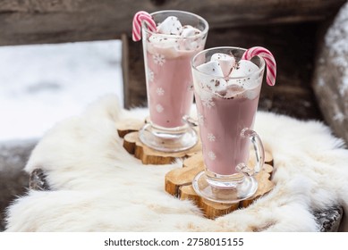 Two glass mugs filled with ruby chocolate hot chocolate topped with whipped cream, marshmallows and a candy cane. The snowflake mugs, wooden coasters and soft fur surface create a cozy, festive winter atmosphere with a distinctive pink ruby chocolate tone.