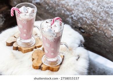 Two glass mugs filled with ruby chocolate hot chocolate topped with whipped cream, marshmallows and a candy cane. The snowflake mugs, wooden coasters and soft fur surface create a cozy, festive winter atmosphere with a distinctive pink ruby chocolate tone.