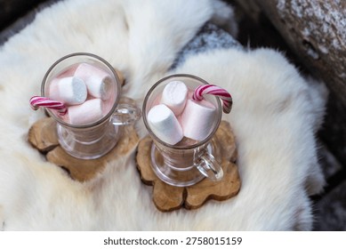 Two glass mugs filled with ruby chocolate hot chocolate topped with whipped cream, marshmallows and a candy cane. The snowflake mugs, wooden coasters and soft fur surface create a cozy, festive winter atmosphere with a distinctive pink ruby chocolate tone.