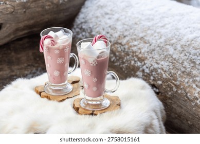 Two glass mugs filled with ruby chocolate hot chocolate topped with whipped cream, marshmallows and a candy cane. The snowflake mugs, wooden coasters and soft fur surface create a cozy, festive winter atmosphere with a distinctive pink ruby chocolate tone.