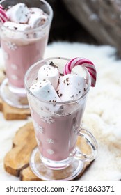 Two glass mugs filled with ruby chocolate hot chocolate topped with whipped cream, marshmallows and a candy cane. The snowflake mugs, wooden coasters and soft fur surface create a cozy, festive winter atmosphere with a distinctive pink ruby chocolate tone.