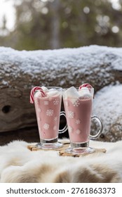 Two glass mugs filled with ruby chocolate hot chocolate topped with whipped cream, marshmallows and a candy cane. The snowflake mugs, wooden coasters and soft fur surface create a cozy, festive winter atmosphere with a distinctive pink ruby chocolate tone.