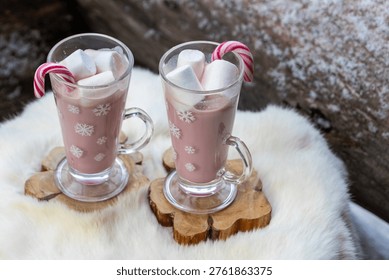 Two glass mugs filled with ruby chocolate hot chocolate topped with whipped cream, marshmallows and a candy cane. The snowflake mugs, wooden coasters and soft fur surface create a cozy, festive winter atmosphere with a distinctive pink ruby chocolate tone.