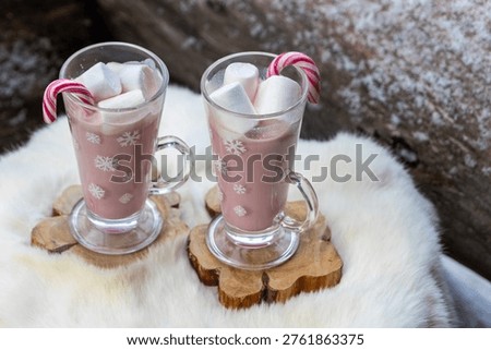 Two glass mugs filled with ruby chocolate hot chocolate topped with whipped cream, marshmallows and a candy cane. The snowflake mugs, wooden coasters and soft fur surface create a cozy, festive winter atmosphere with a distinctive pink ruby chocolate tone.