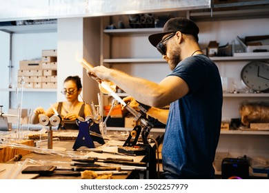 Two glassblowing artisans work in a workshop, one using a torch to heat a glass rod with a vase and the other shaping molten glass on a flame - Powered by Shutterstock