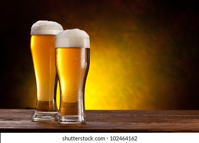 Two Glasses Of Beers On A Wooden Table. Dark Yellow Background.