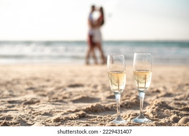 Two glasses with champagne on sand on foreground. Drink wine by seashore. Romantic date of loving couple on seaside. Selective focus. - Powered by Shutterstock