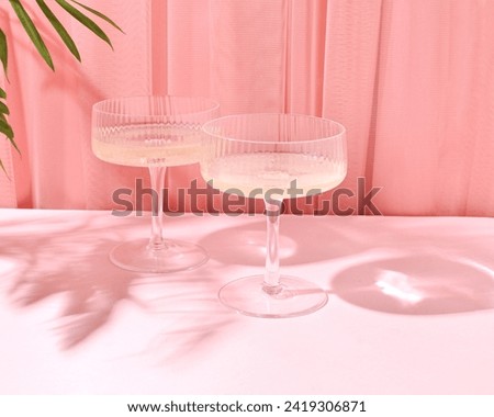 Two glasses of champagne in the shadow of a palm branch, a thin light curtain of pastel peach pink color in the background, retro style, summer party concept.