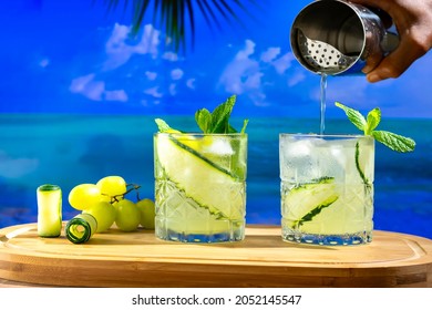 Two Glasses With Drinks On A Wooden Board, In The Background A Beach, Hand Holding A Cocktail Shaker
