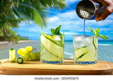 Two Glasses With Drinks On A Wooden Board, In The Background A Beach, Hand Holding A Cocktail Shaker
