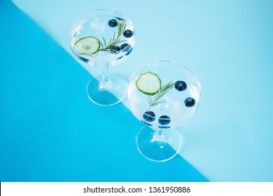 Two Glasses Of A Gin And Tonic Drink With Blueberries, Cucumber And A Rosemary Twig Isolated On Abstract, Colourful, Geometrical Color Block Blue Backgound