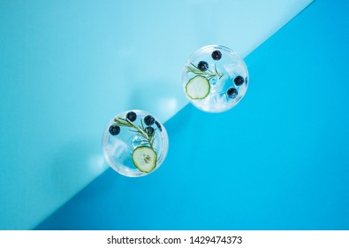 Two glasses of a gin and tonic drink with blueberries, cucumber and a rosemary twig and a yellow lemon isolated on abstract, colourful, geometrical color block blue backgound