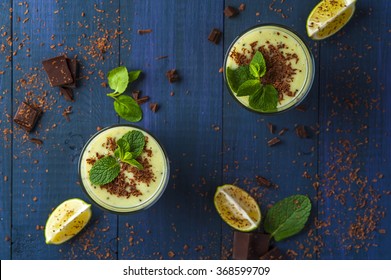 Two glasses of homemade smoothie with kiwi, banana and chocolate chips, mint leaves and pieces of chocolate on blue rustic background. Conception of healthy food. Horizontal view.