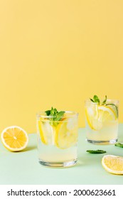 Two Glasses Of Ice Lemon Water With Mint Leaves On A Yellow And Green Background. Fresh Summer Drink With Soda.