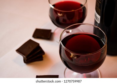 
Two glasses of red wine on a white table next to a dark bottle and pieces of chocolate.