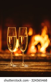 Two Glasses Of Sparkling Champagne In Front Of Warm Fireplace. Cozy Relaxed Magical Atmosphere In A Chalet House By The Fireside. Snug Holiday Concept. Beautiful Background With Shimmering Wine.