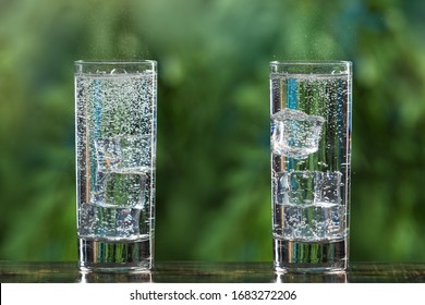 Two Glasses Of Sparkling Water And With Ice Cubes, On A Background Of Greenery, Gas Bubbles And Spray