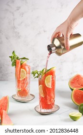 Two Glasses With Tall Long Drink With Watermelon, Lime Slices - And Mint - A Female Hand Pouring Syrup Juice Out Of Cocktail Shaker