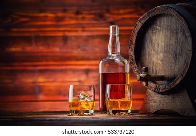 Two Glasses Of Whiskey, Bottle And Keg With Ice Cubes Served On Wooden Planks With Keg. Vintage Countertop With Highlight And A Glass Of Hard Liquor